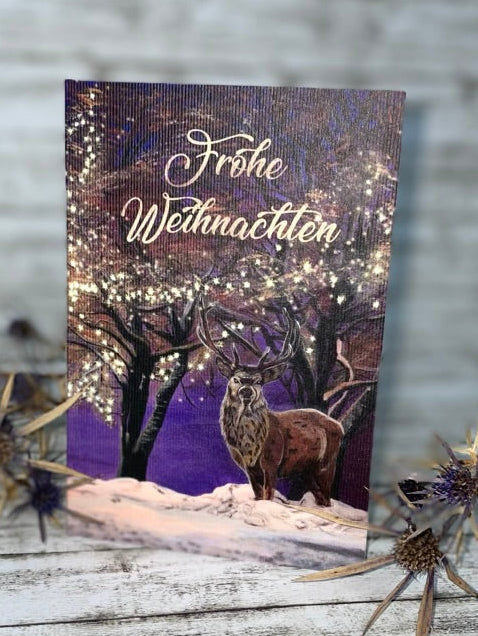 Hirsch unter Lichtern Klappkarte 10x15 cm Acrylgemälde. Ein Hirsch steht auf verschneitem Boden unter dunklen Bäumen, im Hintergrund ein violett-blauer Nachthimmel; winzige Lichter glitzern in den Ästen.