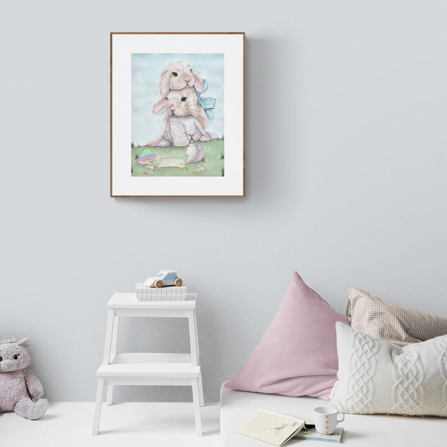 Aquarellgemälde zeigt zwei übereinander sitzende Kaninchen, eines mit blauer Schleife am Ohr, vor zerbrochenem, bunt bemaltem Osterei auf Gras.