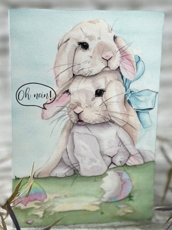Aquarellgemälde zeigt zwei übereinander sitzende Kaninchen, eines mit blauer Schleife am Ohr, vor zerbrochenem, bunt bemaltem Osterei auf Gras.