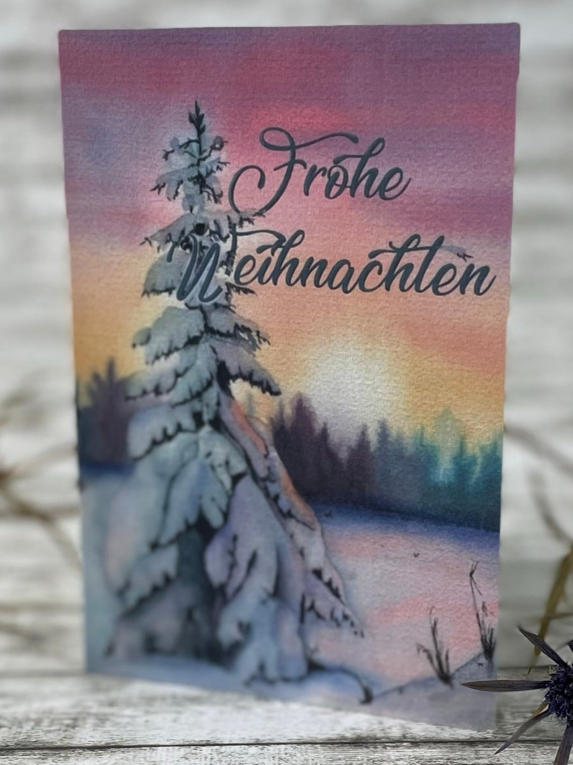 Schneetanne Klappkarte 10x15 cm, Aquarellbild einer verschneiten Winterlandschaft mit einer schneebedeckten Fichte im Vordergrund; sanfter Farbverlauf von rosa, orange und blau am Himmel, Hintergrund mit schmalen Baumreihen; warme Lichtstimmung.